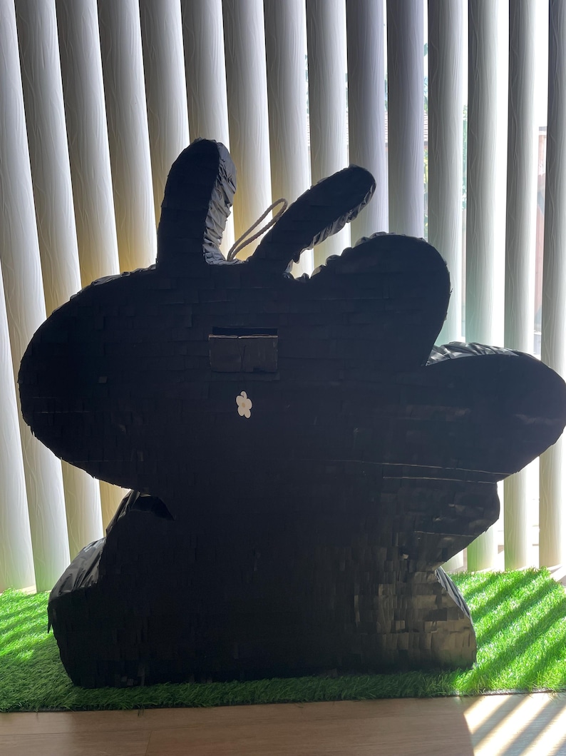 May include: A large, black rabbit-shaped pi&ntilde;ata, crafted from layered paper. It features long ears and a small white flower. The pi&ntilde;ata rests on a patch of green artificial grass.
