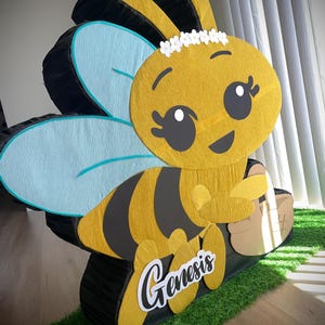 May include: A large, cheerful bee-shaped pi&ntilde;ata. The bee is yellow and black with light blue wings and a white floral crown. The name "Genesis" is written on the side in a cursive font. The pi&ntilde;ata is on a bed of green turf.
