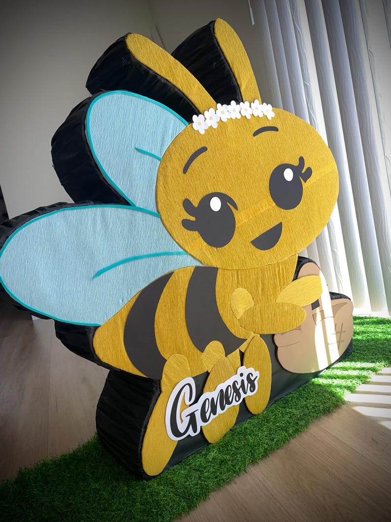 May include: A large, cheerful bee-shaped pi&ntilde;ata. The bee is yellow and black with light blue wings and a white floral crown. The name "Genesis" is written on the side in a cursive font. The pi&ntilde;ata is on a bed of green turf.
