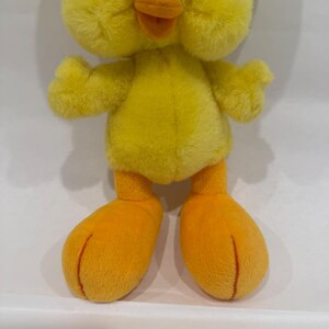 May include: A plush toy shaped like a small yellow chick with soft, fuzzy fur. The figure features a bright yellow body, a rounded head, and two prominent orange feet with visible stitched lines. The beak is a solid, rounded orange color.