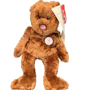 May include: A brown plush teddy bear with a round nose and small black eyes. The bear has a small pin with the American flag on its nose and a 2022 FIFA pin on its chest. A tag is attached to the bear.