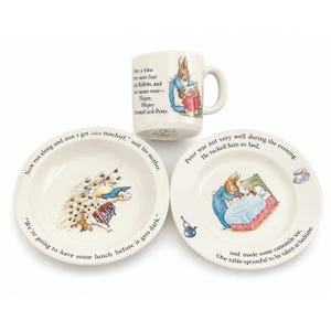 May include: A ceramic three-piece dinnerware set featuring Peter Rabbit illustrations and text. The set includes a mug, a bowl, and a plate, all in cream-coloured ceramic with colourful character graphics and printed storybook text in black lettering.