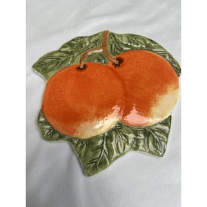 Puede incluir: Un plato decorativo de cerámica con dos frutas de color naranja con tallos marrones, sobre una base verde en forma de hoja. El plato tiene un acabado brillante y probablemente está pintado a mano, mostrando un diseño con temática de frutas.
