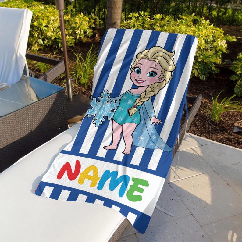 K&ouml;nnte beinhalten: Blau-wei&szlig; gestreiftes Strandtuch mit einer Cartoon-Illustration einer Figur, die eine Schneeflocke h&auml;lt. Das Handtuch hat das Wort "NAME" in bunten Buchstaben am unteren Rand. Das Handtuch liegt auf einer wei&szlig;en Liege.