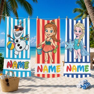 K&ouml;nnte beinhalten: Drei Strandt&uuml;cher h&auml;ngen an einer W&auml;scheleine. Jedes Handtuch zeigt eine Cartoonfigur: Olaf der Schneemann, ein M&auml;dchen mit Zopf und Elsa. Die Handt&uuml;cher haben farbige Streifen und das Wort "NAME" unten.