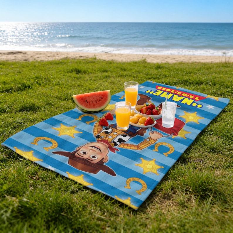 May include: A blue and white striped beach blanket featuring a cartoon cowboy character, stars, and horseshoes. The blanket is set up on a grassy area with a beach and ocean in the background. There are drinks and snacks on the blanket.