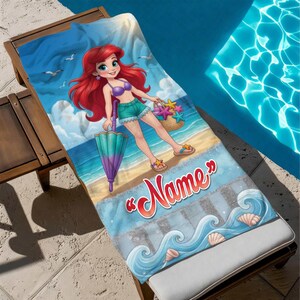 Op de afbeelding: Een strandlaken met een cartoonillustratie van een zeemeermin op een strand. De zeemeermin heeft rood haar en houdt een paraplu en een mand met zeesterren vast. De handdoek heeft het woord "Name" in rood script en een blauw oceaan golf ontwerp.