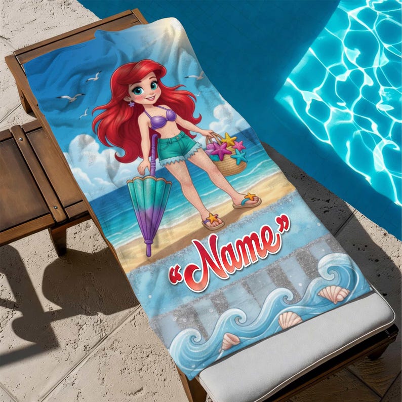 Op de afbeelding: Een strandlaken met een cartoonillustratie van een zeemeermin op een strand. De zeemeermin heeft rood haar en houdt een paraplu en een mand met zeesterren vast. De handdoek heeft het woord "Name" in rood script en een blauw oceaan golf ontwerp.
