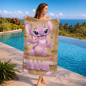 May include: A pink beach towel featuring the character Angel from Lilo & Stitch. The towel has a sand-colored background with hearts and seashells. The word "NAME" is printed in pink block letters.