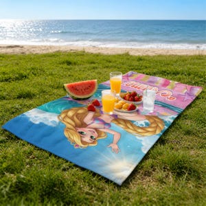 Op de afbeelding: Een strandlaken met een cartoon illustratie van een prinses met lang blond haar, op een blauw en roze golfpatroon. Het laken ligt op gras, met een strand en zee op de achtergrond. Er is watermeloen, fruit en drankjes.