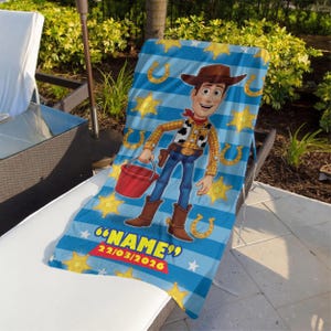 May include: A blue and white striped beach towel featuring a cartoon image of Woody from Toy Story. The towel has yellow stars and horseshoes, with the text "NAME" and a date. The towel is draped over a lounge chair.