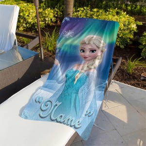 May include: A blue beach towel featuring a cartoon image of a blonde woman in a blue dress. The towel has the word "Name" in a cursive font. The background has a blue and purple aurora borealis design with snowflakes.