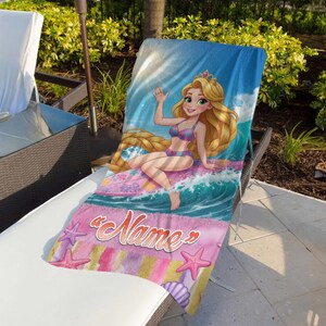 Op de afbeelding: Een strandlaken met een cartoon illustratie van een prinses. De prinses heeft lang blond haar en draagt een bikini, zittend op een golf. De handdoek heeft een blauwe hemel achtergrond en een roze gedeelte met het woord "Name" en zeesterren.