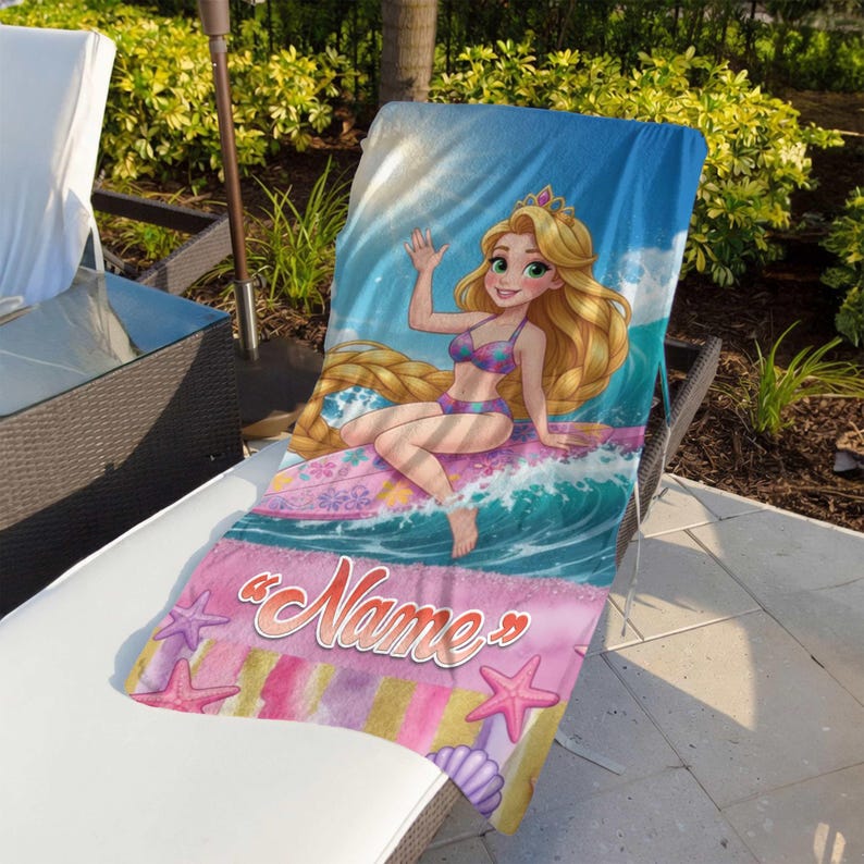 Op de afbeelding: Een strandlaken met een cartoon illustratie van een prinses. De prinses heeft lang blond haar en draagt een bikini, zittend op een golf. De handdoek heeft een blauwe hemel achtergrond en een roze gedeelte met het woord "Name" en zeesterren.