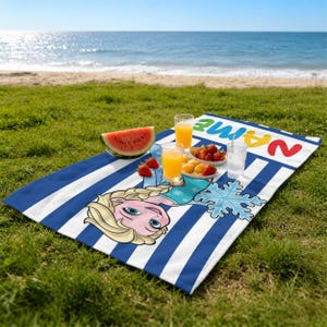 K&ouml;nnte beinhalten: Ein blau-wei&szlig; gestreiftes Strandtuch mit einem Cartoon-Bild von Elsa aus Frozen, einer Schneeflocke und dem Wort "NAME" in bunten Buchstaben. Das Handtuch liegt auf gr&uuml;nem Gras mit einem Strand und Ozean im Hintergrund. Es gibt eine Wassermelone, Obst und Getr&auml;nke auf dem Handtuch.