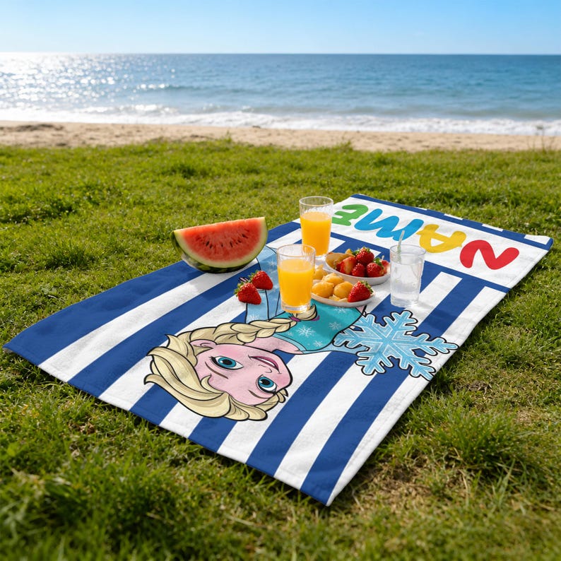 K&ouml;nnte beinhalten: Ein blau-wei&szlig; gestreiftes Strandtuch mit einem Cartoon-Bild von Elsa aus Frozen, einer Schneeflocke und dem Wort "NAME" in bunten Buchstaben. Das Handtuch liegt auf gr&uuml;nem Gras mit einem Strand und Ozean im Hintergrund. Es gibt eine Wassermelone, Obst und Getr&auml;nke auf dem Handtuch.