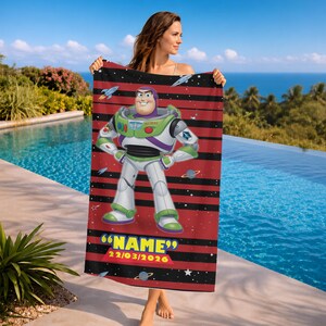 May include: A red and black striped beach towel featuring a cartoon image of Buzz Lightyear. The towel has the text "NAME" and the date 22/03/2026. The towel is held up by a person near a pool.