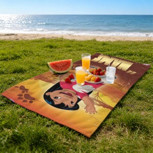 May include: A beach towel featuring a cartoon character in a red and white outfit, lying on a grassy area near the beach. The towel has a brown border with gold text. There is a watermelon slice, fruit, and drinks on the towel.