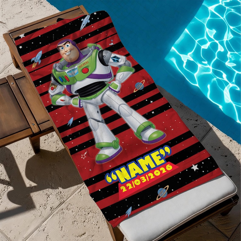 May include: A red and black striped beach towel featuring Buzz Lightyear from Toy Story. The towel has a personalized name and date in yellow text. The towel is on a wooden lounge chair next to a pool.