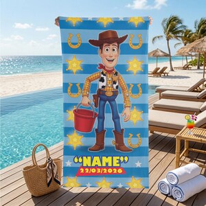 May include: A blue and white striped beach towel featuring a cartoon image of Woody from Toy Story. The towel has yellow stars and horseshoes, with the name "NAME" and the date 22/03/2026 printed at the bottom. A red bucket is in Woody's hand.