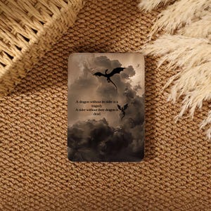 Könnte beinhalten: Ein rechteckiger Untersetzer mit einem dunklen, dramatischen Bild von Drachen, die in einem bewölkten Himmel fliegen. Der Untersetzer trägt den Text: "Ein Drache ohne seinen Reiter ist eine Tragödie. Ein Reiter ohne seinen Drachen ist tot."