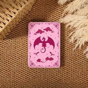 May include: A pink rectangular card featuring a dark magenta dragon with outstretched wings, clouds, and smaller dragons. The card has a decorative border and is set against a textured brown surface.