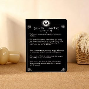Könnte beinhalten: Ein schwarzes, rechteckiges Notizbuch mit dem weißen Text Death Note How to Use It I, einem Totenkopf-Symbol und fünf nummerierten Regeln zur Verwendung des Notizbuchs. Das Cover-Design enthält dekorative weiße Eckverzierungen und weißen Text.