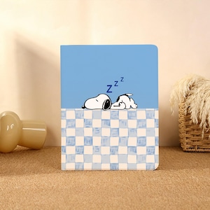 Pode incluir: Um caderno azul com uma ilustração de Snoopy a dormir, com a letra "Z" acima da sua cabeça. A metade inferior tem um padrão xadrez azul e branco. O caderno é retangular e fechado.
