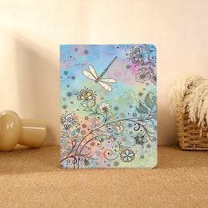 May include: A colorful journal with a dragonfly and floral design. The cover features a watercolor background with a dragonfly and floral illustrations in black and white. The journal is standing upright on a neutral surface.