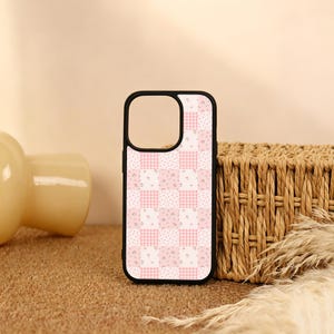 Peut inclure: Une coque de téléphone noire avec un motif à carreaux alternant des carrés vichy rose clair et des carrés blancs ornés de petites fleurs roses. La coque présente une grande découpe carrée en haut pour le bloc de l'objectif de l'appareil photo.