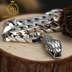 May include: A silver bracelet with a snake head design. The bracelet is made of interlocking links and features a detailed snake head with open jaws. The bracelet is displayed on a wooden surface.