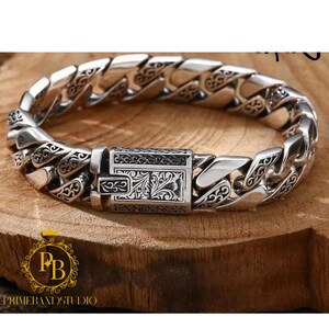 May include: A silver-toned metal chain link bracelet featuring intricate engraved floral and scroll patterns on each individual link and a rectangular box clasp closure with matching detailed engravings on the front surface.