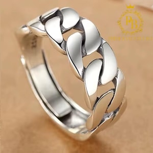 Puede incluir: Un anillo plateado con un diseño de cadena de eslabones. El anillo presenta una serie de eslabones ovalados aplanados e interconectados, creando una apariencia audaz y texturizada. El anillo es ajustable y tiene un acabado pulido, que refleja la luz.