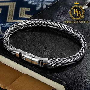Op de afbeelding: Een zilverkleurige tarwekettingarmband met een gepolijste metalen baksluiting. De armband heeft een gevlochten, getextureerd schakelontwerp met donkere verdiepte gebieden tussen de schakels, wat zorgt voor een contrastrijk uiterlijk over de hele ketting.