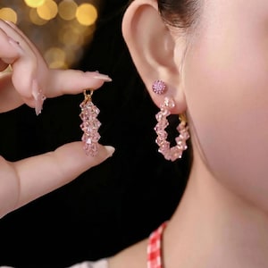 May include: A pair of hoop earrings featuring light pink faceted crystals arranged in a circular pattern. One earring is worn on the earlobe, while the other is held up to show the design. A small pink stud earring is also visible on the ear.