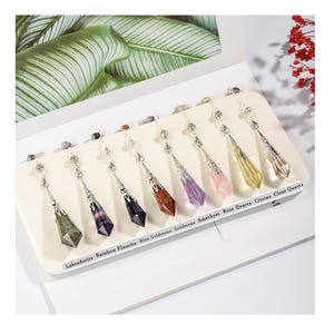 May include: A set of eight faceted crystal pendulums hanging from silver-toned metal caps and chains. The stones are labeled Labradorite, Rainbow Fluorite, Blue Goldstone, Goldstone, Amethyst, Rose Quartz, Citrine, and Clear Quartz on the display.