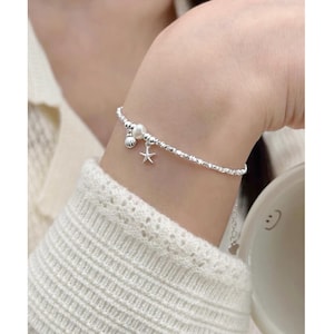 May include: A silver beaded bracelet featuring a small white pearl, a tiny silver seashell charm, and a delicate silver starfish charm. The bracelet is constructed from small, textured metallic beads that create a shimmering effect along the chain.