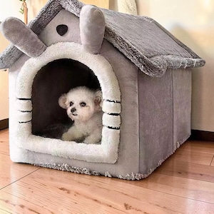 以下が含まれることがあります： ウサギの耳の形をした屋根を持つ灰色のぬいぐるみペットハウス。アーチ型の入り口の周りには柔らかくふわふわした白いトリムがあり、小さな黒い水平線で装飾されています。小さな白い犬が囲いの中に座っています。