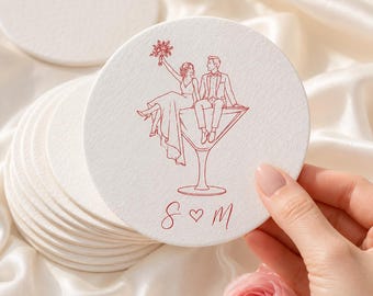 May include: White round coasters with a red line drawing of a couple in a martini glass. The initials "S ♡ M" are below the drawing. A stack of coasters is visible, with a pink rose in the foreground.