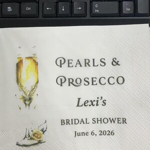 May include: A white paper napkin featuring a watercolor illustration of a champagne flute with pearls and a yellow rose. The text reads: Pearls & Prosecco, Lexi's BRIDAL SHOWER, June 6, 2026. A metal ruler is visible at the bottom showing 30 cm.