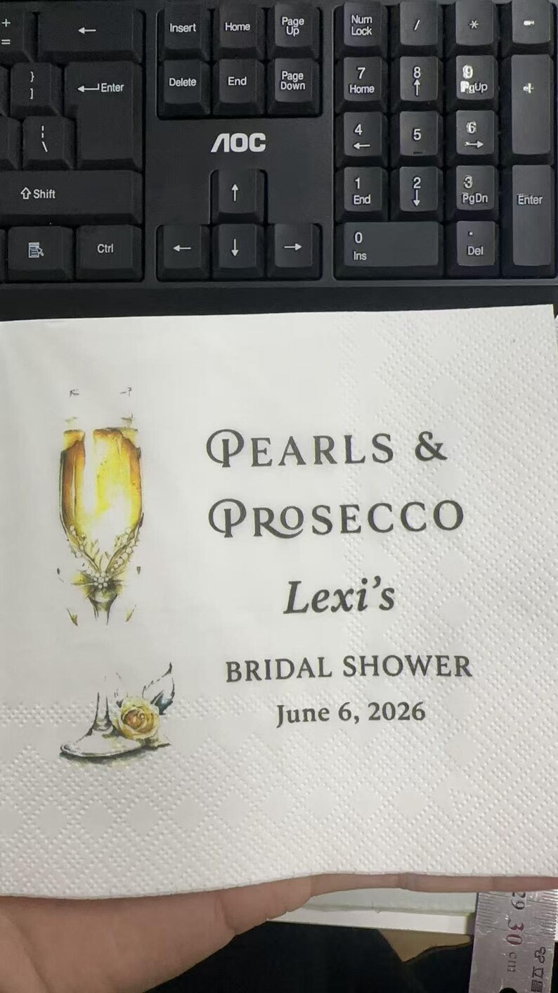 May include: A white paper napkin featuring a watercolor illustration of a champagne flute with pearls and a yellow rose. The text reads: Pearls & Prosecco, Lexi's BRIDAL SHOWER, June 6, 2026. A metal ruler is visible at the bottom showing 30 cm.