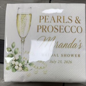 May include: A white paper napkin featuring a gold-toned illustration of a champagne flute filled with bubbly liquid. The text reads Pearls & Prosecco, Miranda's Bridal Shower, and July 25, 2026, accompanied by green leaves and white pearls.