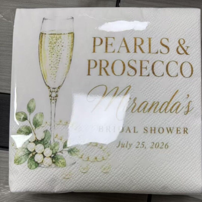 May include: A white paper napkin featuring a gold-toned illustration of a champagne flute filled with bubbly liquid. The text reads Pearls & Prosecco, Miranda's Bridal Shower, and July 25, 2026, accompanied by green leaves and white pearls.