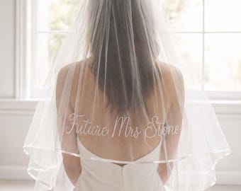 May include: A bride wearing a white wedding veil with the text "Future Mrs Stone" embroidered on the back. The veil is sheer and layered, with a satin ribbon trim. The bride is wearing a white dress.