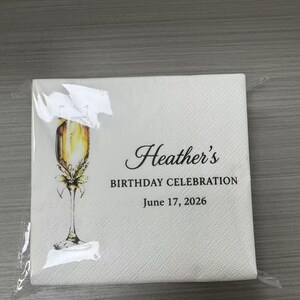 May include: A square white paper napkin featuring a printed illustration of a champagne flute filled with golden liquid. The text reads Heather's BIRTHDAY CELEBRATION June 17, 2026 in black lettering. The napkin is sealed in clear plastic.