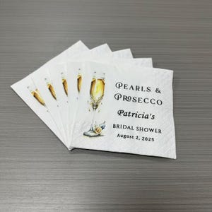 May include: A stack of white paper napkins featuring a watercolor illustration of a champagne flute with a pearl accent. The text reads: Pearls & Prosecco, Patricia's, BRIDAL SHOWER, August 2, 2025, printed in black serif and sans-serif fonts.