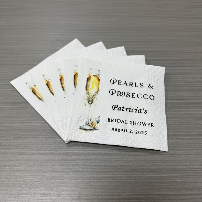May include: A stack of white paper napkins featuring a watercolor illustration of a champagne flute with a pearl accent. The text reads: Pearls & Prosecco, Patricia's, BRIDAL SHOWER, August 2, 2025, printed in black serif and sans-serif fonts.