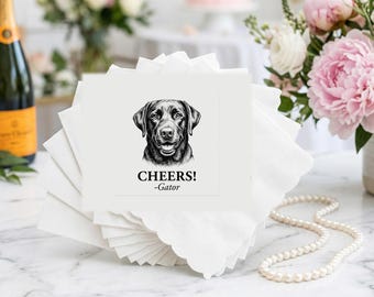 May include: A stack of white paper napkins with scalloped edges, featuring a black and white illustration of a Labrador retriever dog head. Below the dog, the text reads CHEERS! and the name Gator in a black serif font centered on the napkin.