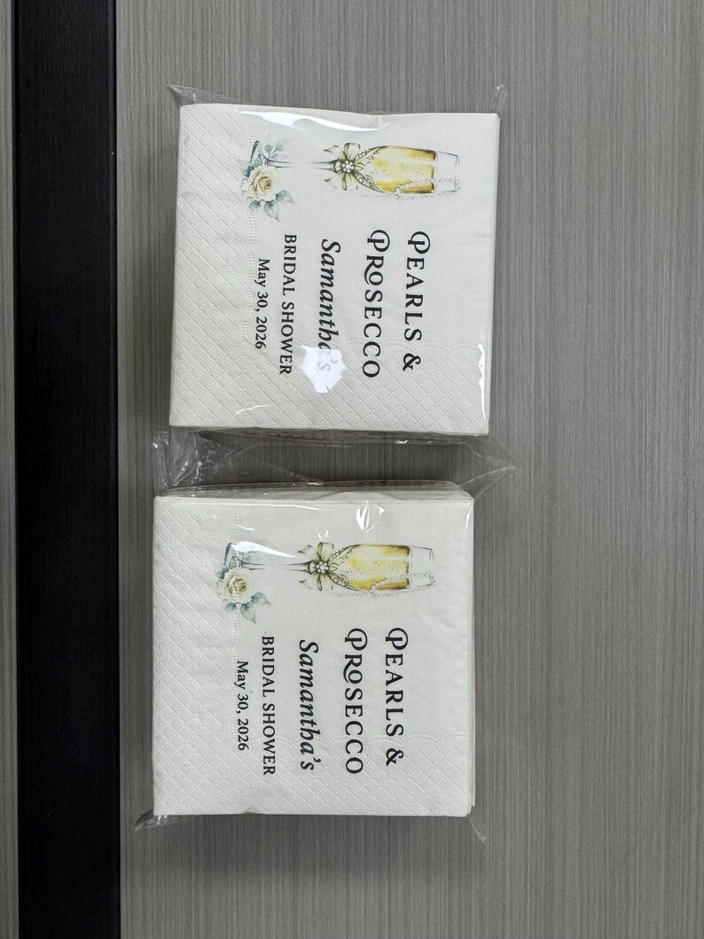May include: Two stacks of white paper napkins in clear plastic packaging. Each napkin features a printed illustration of a champagne flute with a white rose and the text: PEARLS & PROSECCO, Samantha's, BRIDAL SHOWER, May 30, 2026.