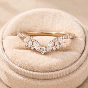 Könnte beinhalten: Ein goldfarbener Ring mit geschwungenem Design. Der Ring hat eine zentrale Anordnung von Steinen im Marquiseschliff und einen einzelnen runden Stein, mit kleineren Steinen entlang des Bandes. Der Ring ist vor einem beigen Hintergrund platziert.
